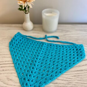 A turquoise crocheted bandana lays on a dresser. This boho style hair scarf has a delicate open stitch pattern. There are ties on the bandana to secure to the hair and head.