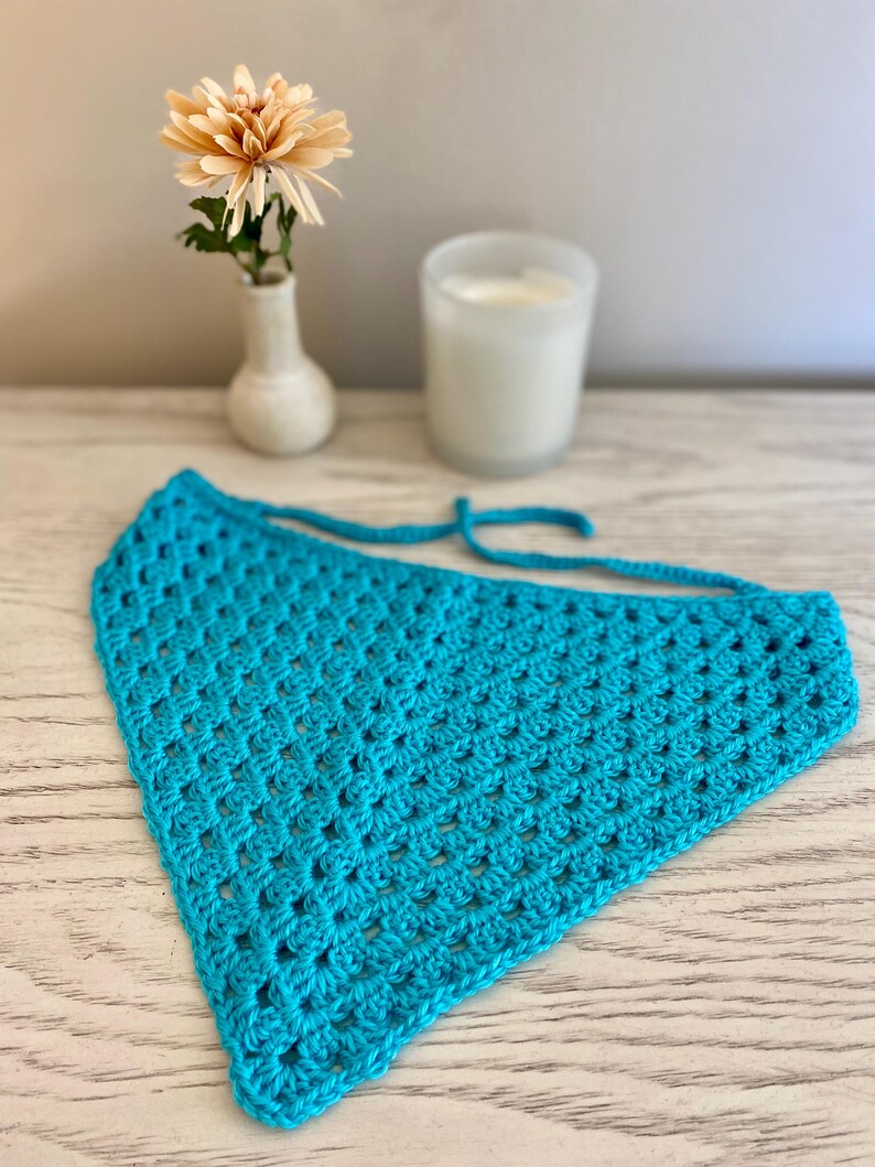A turquoise crocheted bandana lays on a dresser. This boho style hair scarf has a delicate open stitch pattern. There are ties on the bandana to secure to the hair and head.