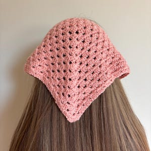 May include: A dusty rose triangular headscarf crafted with a crochet granny square pattern. The accessory features a textured, open-work stitch design that forms a point at the center, covering the top portion of the head with a soft pink hue.