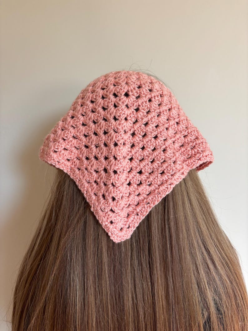 May include: A dusty rose triangular headscarf crafted with a crochet granny square pattern. The accessory features a textured, open-work stitch design that forms a point at the center, covering the top portion of the head with a soft pink hue.