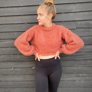 May include: A woman wearing a cropped, fuzzy, rust-colored sweater with long sleeves and black leggings.