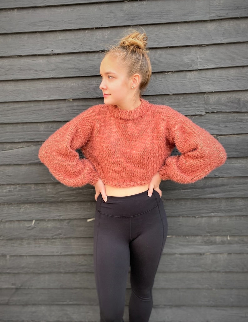 May include: A woman wearing a cropped, fuzzy, rust-colored sweater with long sleeves and black leggings.