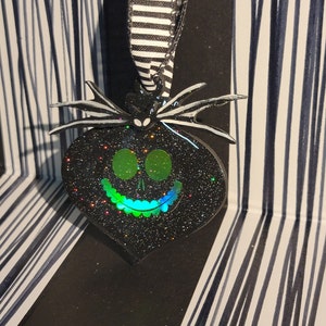 Pode incluir: Adorno de coração preto e verde com glitter com um rosto inspirado em Jack Skellington. O adorno tem uma fita listrada preta e branca presa ao topo.