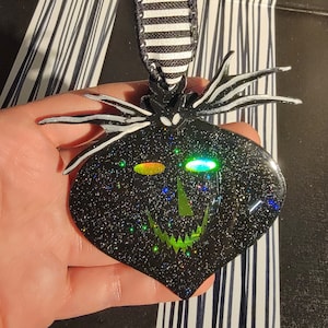 Puede incluir: Adorno navideño de purpurina negro y verde con forma de calabaza con una sonrisa de dientes y ojos iridiscentes.