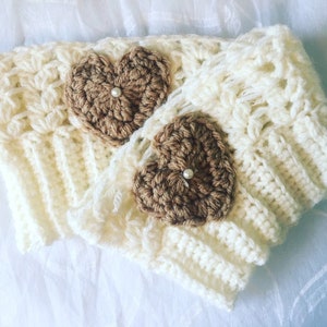 Puede incluir: Calentadores de piernas de ganchillo color crema con dos corazones de ganchillo marrón, cada uno adornado con una pequeña perla. Los calentadores de piernas presentan un patrón de puntada texturizada y se muestran sobre una superficie blanca.