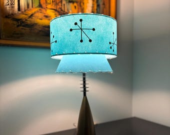 May include: A mid-century modern table lamp with a teal shade featuring a black starburst pattern. The lamp has a brass-colored conical base and a black circular base. The lamp is on a dark surface.