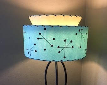 May include: A mid-century modern table lamp with a blue and white shade featuring a black starburst pattern. The lamp has a black metal base with a curved design.