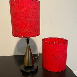 May include: Two red lampshades with black stitching and a black and gold lamp base. The lampshades are made of a textured fabric.