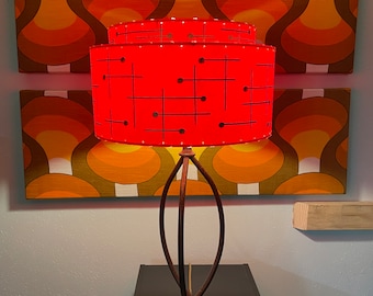 May include: A red lampshade with a geometric pattern sits atop a black metal lamp base. The lamp is on a black table with a wood shelf behind it. The lamp is in front of a wall with a retro patterned wallpaper.