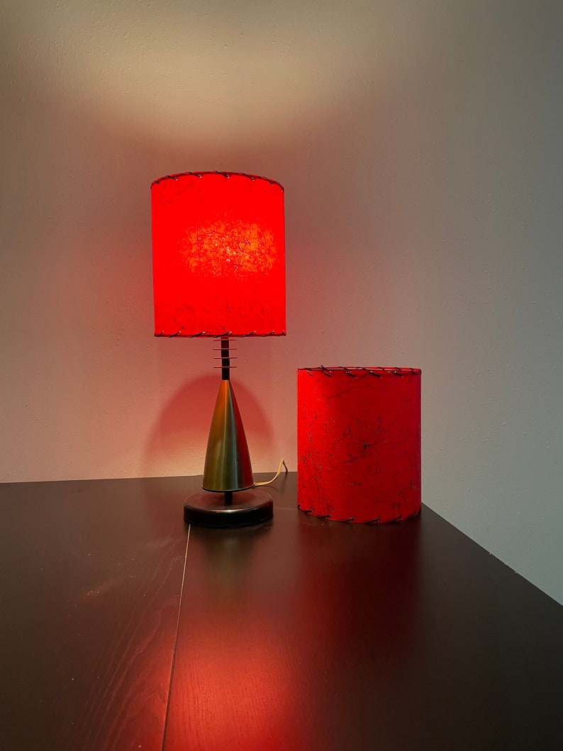 May include: Two red lampshades with a textured fabric surface. The lampshade on the left is on a gold metal lamp base with a black base. The lampshade on the right is sitting on a dark wood surface.