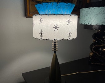 May include: A mid-century modern table lamp with a white and black star patterned lampshade and a blue and gold top lampshade. The lamp has a gold base and a black base.