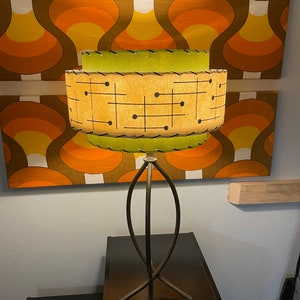 Puede incluir: Una lámpara de mesa moderna de mediados de siglo con una pantalla de patrón geométrico verde y beige. La lámpara tiene una base de metal negro con un diseño curvo único. La lámpara está sentada en una mesita de noche negra.