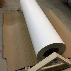 May include: A large roll of white paper, likely for printing or art projects, is supported by a wooden stand. The paper is a bright white color and appears to be smooth. The stand is made of light-colored wood and is positioned on a piece of cardboard.