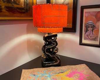 May include: A black ceramic table lamp with a gold snake design and an orange geometric patterned lampshade. The lamp is on a table with a brown paper with a colorful painting of two birds.