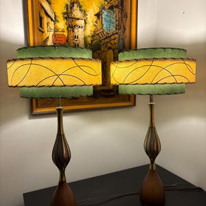 Puede incluir: Par de lámparas de mesa de mediados de siglo. Cada lámpara tiene una base de madera, un centro de metal color latón y una pantalla de dos niveles. Las pantallas son amarillas con un diseño de remolino negro y ribete verde. Una pintura vintage cuelga en el fondo.