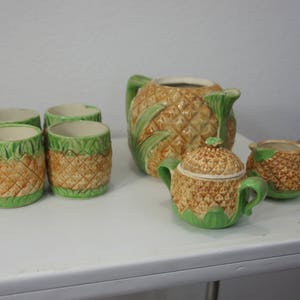 Könnte beinhalten: Ein Set aus Keramik-Teetassen und einer Teekanne im Ananas-Design. Die Teekanne hat ein grünes und braunes Ananas-Muster. Die Teetassen sind kleiner und haben ein ähnliches Design.