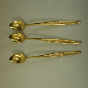 May include: Three gold-colored spoons with ornate designs on the handles. The spoons are arranged in a row, with the handles pointing to the right.