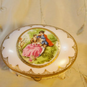 May include: Oval porcelain trinket box with a gold trim and a painted scene of a couple in a garden. The box has a hinged lid and sits on three small feet.