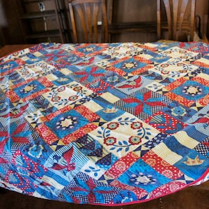 May include: A round, white tablecloth with a patchwork design featuring red, blue, and white squares and floral patterns. The tablecloth is made of cotton and has a fringed edge.