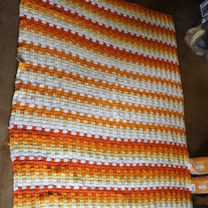 Op de afbeelding: Een gehaakt vloerkleed met oranje, witte en gele strepen. Het kleed is gemaakt met een eenvoudig steekpatroon en heeft een textuur.