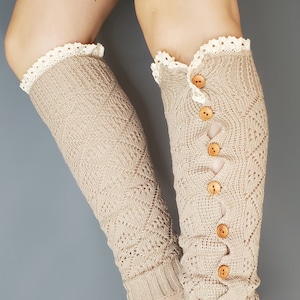 Op de afbeelding: Beige gebreide beenwarmers met een kanten rand aan de bovenkant en houten knopen aan de zijkant. De beenwarmers hebben een ruitpatroon en worden tegen een grijze achtergrond getoond.