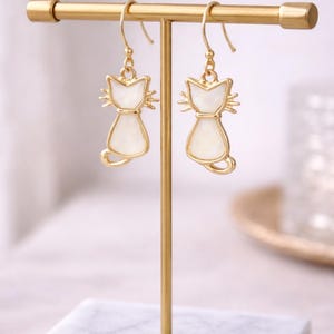 May include: Gold-tone cat-shaped earrings with a white enamel fill, displayed on a gold jewelry stand with a marble base. The earrings feature a simple cat silhouette with a curved tail and whiskers.