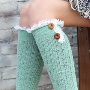 May include: Mint green knee-high socks with a delicate openwork pattern. The top of each sock is trimmed with white lace and accented with two brown buttons. The socks are shown against a brick wall and a brown suitcase.