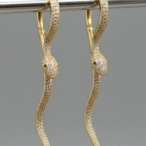 May include: A pair of gold-colored snake earrings. Each earring features a serpentine design encrusted with small, sparkling stones. The snake's head has a small green stone for the eye. The earrings are suspended from a gold-colored hoop.