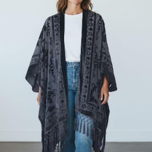 May include: A dark grey kimono-style wrap with a black floral pattern and fringe trim. The wrap is worn over a white top and blue jeans. The kimono has long sleeves and a relaxed fit, suitable for layering.