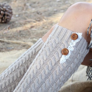May include: Knit leg warmers in a light beige color with a cable knit design. The leg warmers have two brown buttons and white lace trim. A fringed scarf is visible in the background.