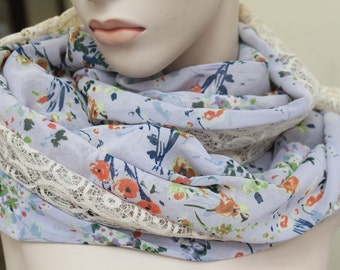 May include: A light blue infinity scarf with a floral pattern in orange, green, and blue. The scarf is trimmed with a cream-colored lace. The scarf is wrapped around a mannequin's neck.