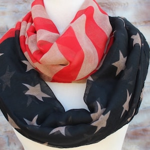 Puede incluir: Bufanda infinita con diseño de la bandera estadounidense. Tiene rayas rojas y blancas y una sección negra con estrellas color beige. La bufanda se muestra en un maniquí. Un accesorio elegante para cualquier guardarropa.