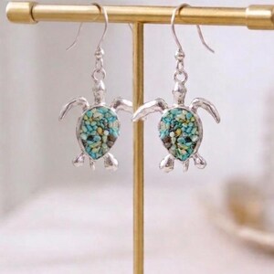 May include: Silver-toned turtle earrings with turquoise and yellow stone inlay. The earrings are displayed on a gold-colored jewelry stand. The turtle design features outstretched flippers and a detailed shell.