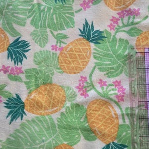 May include: Cream-colored fabric with a tropical pattern featuring yellow pineapples, green monstera leaves, and small pink flowers. The pineapples have green spiky tops. The design is repeated across the fabric.