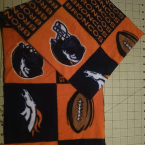 May include: Orange and navy blue fleece scarf featuring a repeating pattern of football helmets, footballs, and the word "BRONCOS". The scarf has a fringed edge and is folded over.