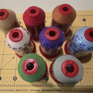 May include: A collection of seven spools of thread in various colors, including beige, red, orange, purple, blue, green, and gray. Each spool has a red top and is arranged on a yellow cutting mat.