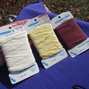 May include: Three spools of Wicking Yarn in cream, yellow, and burgundy, each labeled with "50 YARDS" and "Cross Stitch Originals." The spools are displayed against a blue fabric background.