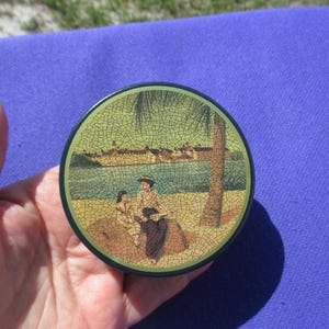 May include: Round, vintage tin with a painted scene of people on a beach with a palm tree and buildings in the background. The tin has a green border and a dark blue rim.