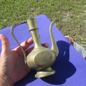 May include: A brass pitcher featuring an elongated neck, a curved handle, and a long, slender spout. The surface of the metal is decorated with intricate, etched floral patterns and leaf motifs that wrap around the body, neck, and spout.