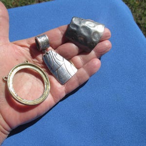 May include: A collection of silver and gold-toned jewelry components. Includes a rectangular pendant with a textured design, a hammered metal square, and a gold-toned circular frame. The items are displayed on a blue surface.