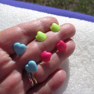 May include: A collection of colorful, small, heart-shaped and seashell-shaped charms. The charms are in shades of light blue, neon green, and hot pink. The heart charms are on a gold ring.