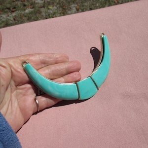 May include: A turquoise-colored necklace with a crescent shape. The necklace has a gold-colored metal trim and a marbled effect on the turquoise sections. The necklace is held in a hand.