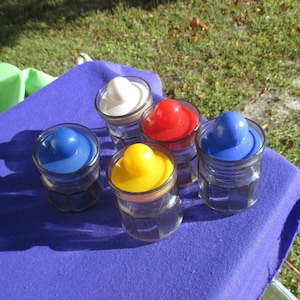 May include: Five small glass jars with colorful lids in white, red, yellow, and two shades of blue. The jars are arranged on a purple surface, with a grassy background visible. The lids are rounded and sit atop the clear glass jars.