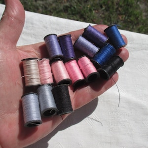 May include: A collection of spools of thread in various colors, including pink, blue, and gray. The spools are arranged in a hand, with some spools stacked on top of others.