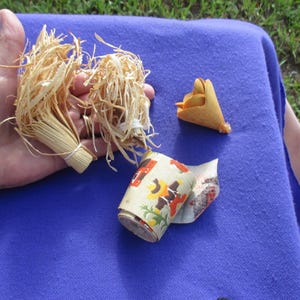 May include: A collection of small, handmade items. Two bundles of dried, light-colored straw are held together with twine. A small, folded, cone-shaped object and a rolled paper with a floral pattern are also visible.