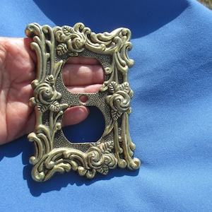May include: Ornate brass-colored electrical outlet cover with a floral rose design. The cover features two circular openings for the outlets and is surrounded by detailed, sculpted roses and leaves. The background is a solid blue fabric.