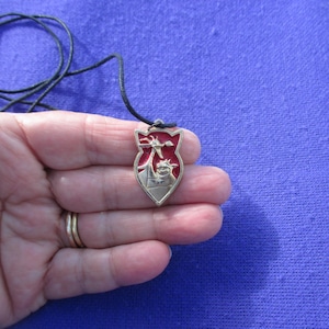 May include: A shield-shaped pendant featuring a metallic gold-toned dragon figure set against a vibrant red enamel background. The pendant is suspended from a thin black cord, creating a necklace with a distinct heraldic design and clear metal edges.