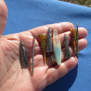 May include: A collection of small, decorative pendants and charms in various shapes and materials. Includes silver-toned figures, twisted metal rods, and colorful, translucent pieces. The items are held in a hand against a blue background.