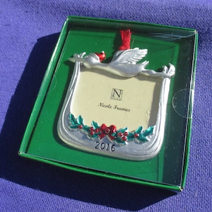 May include: A silver-colored Christmas ornament in a green box. The ornament is shaped like a picture frame with a flying bird at the top. It has a red ribbon for hanging and is decorated with holly and the year 2016. The text "Nicole Frames" is visible.