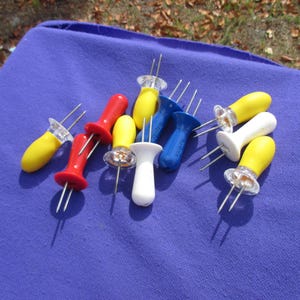 May include: A collection of colorful corn holders in red, yellow, white, and blue. Each holder has a plastic handle and two metal prongs for securing corn on the cob. The holders are arranged on a blue surface.